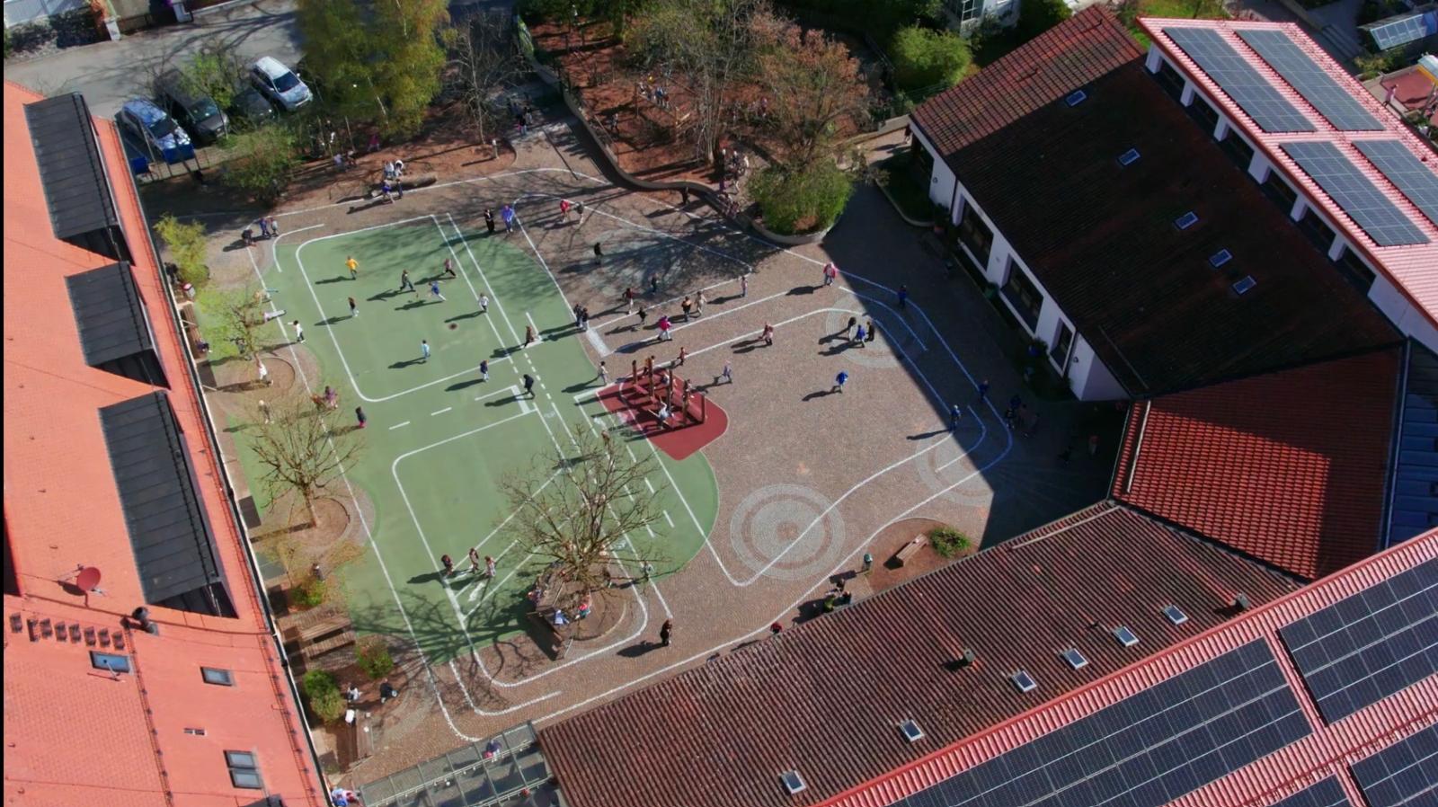 Eine spektakuläre Drohnen-Aufnahme aus der Vogelperspektive zeigt den bunt bemalten Schulhof der Grundschule Pullach mit Laufspuren und spielenden Kindern an der frischen Luft.