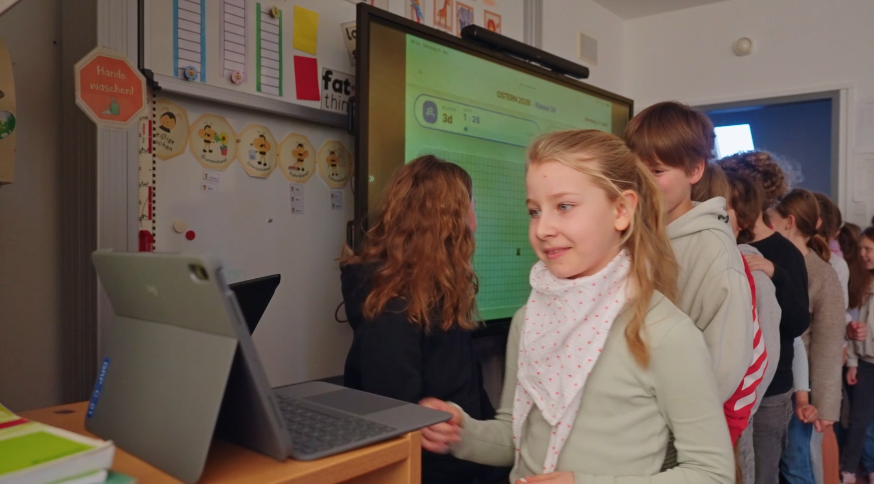 Fröhliche Grundschulkinder stehen im Klassenzimmer geduldig in einer Reihe an, um morgens spielerisch und eigenständig am Tablet ihren autofreien Schulweg im Morgenkreis zu erfassen.